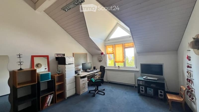A cozy attic room with slanted ceilings, large windows with orange curtains, a desk with a computer, a chair, shelves, and a radiator.