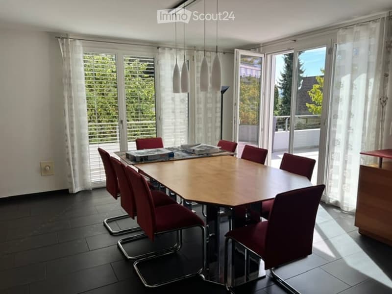 Bright dining room with large windows and a view, featuring a wooden dining table surrounded by red chairs, pendant lights, and a cabinet.