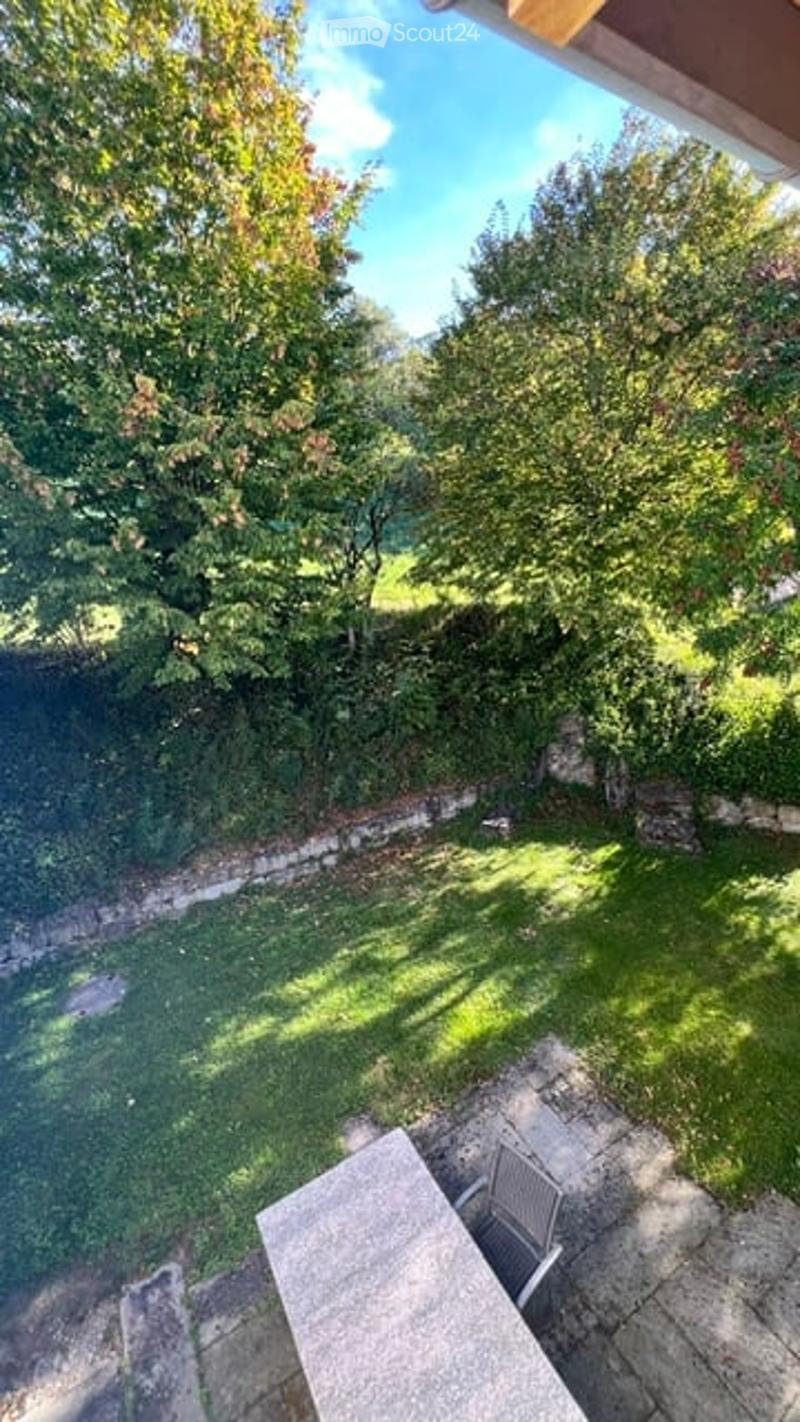 A well-maintained garden with lush green trees, a stone wall, and a patio area with a table and chairs.