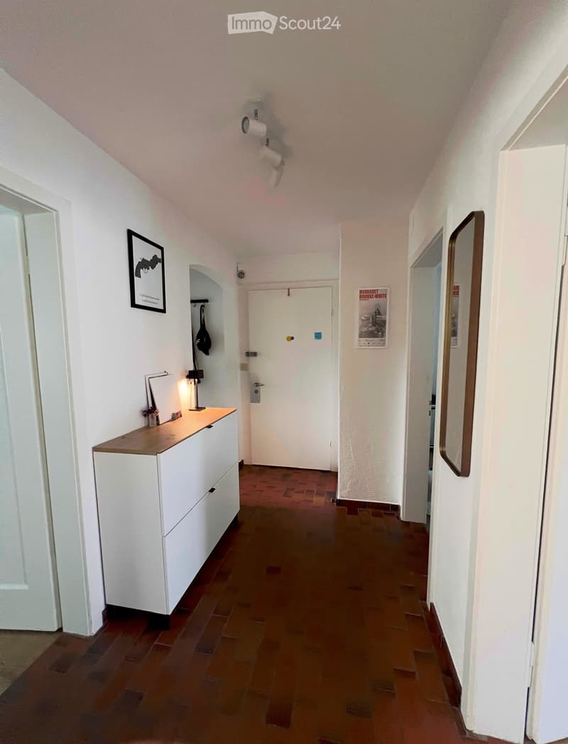 A narrow hallway with white walls, wooden tiled flooring, and several doors. A small cabinet with a lamp and a framed picture is visible on the left side.
