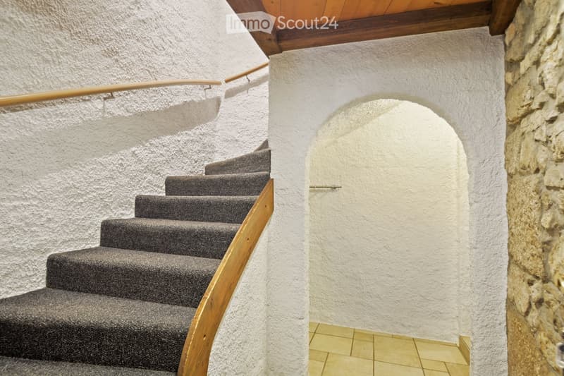 Staircase with wooden handrail and gray carpeted steps, white textured walls, and a stone wall on the right side.