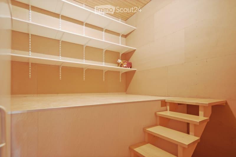 Empty storage room with wooden shelves and a small staircase leading to an upper level. The walls are painted a warm, light orange color, and the ceiling is made of light wood panels.
