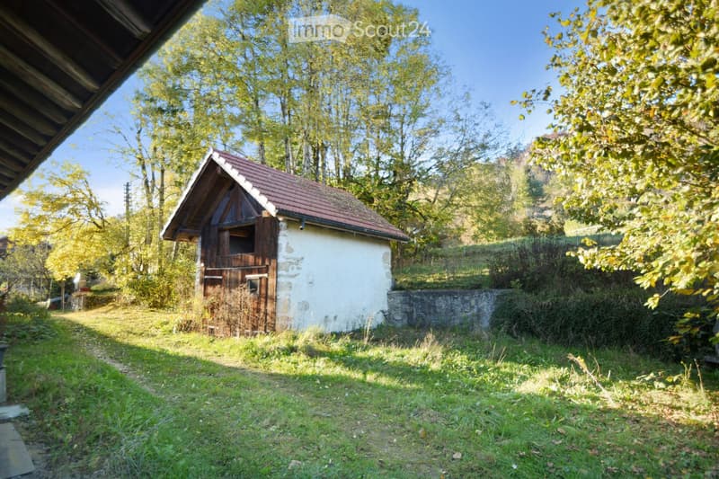 A small, rustic wooden building with a red-tiled roof, surrounded by trees and greenery in a serene outdoor setting.