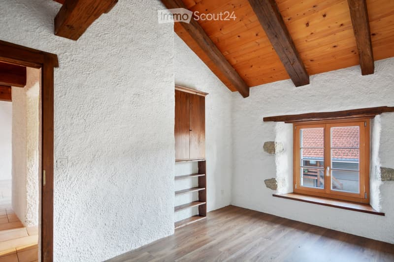 Empty room with exposed wooden beams, white textured walls, wooden floor, built-in wooden shelving unit, and a window with wooden frame showing a view of a roof.