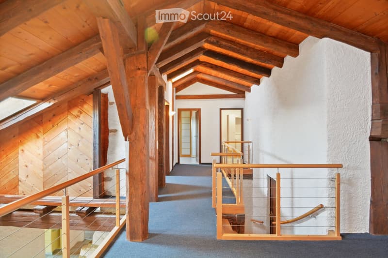 Spacious hallway with exposed wooden beams and a high, slanted wooden ceiling. Features a wooden staircase with a modern glass railing and a blue carpeted floor. The walls are white, and there are several doors leading to other rooms.
