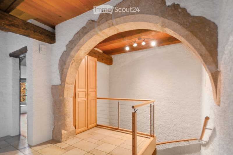A rustic hallway with exposed wooden beams, stone walls, and a wooden door leading to another room. The hallway features a wooden railing and warm lighting.