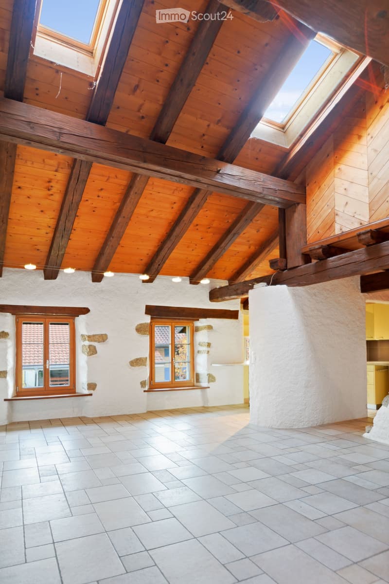 Empty room with exposed wooden beams, white tiled floor, and large windows with wooden frames. The walls are white with stone accents, and the ceiling has skylights.