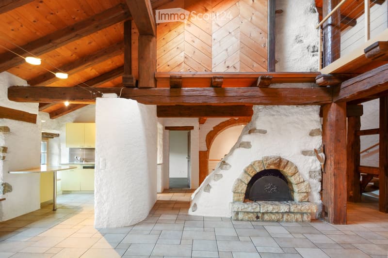 A rustic living space with exposed wooden beams, a stone fireplace, a small kitchen area with cabinets, and a table.