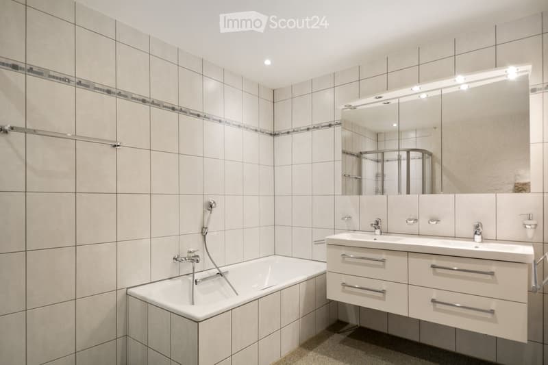 Modern bathroom with a bathtub, double sink, large mirror with integrated lighting, and tiled walls.