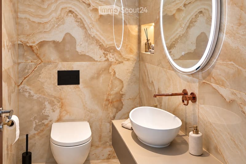 Modern bathroom with marble walls, a white toilet, a sleek sink with a modern faucet, a round illuminated mirror, and a soap dispenser on the counter.