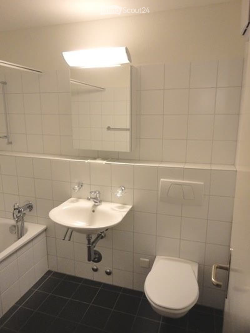 Modern bathroom with white tiled walls, black tiled floor, a sink, toilet, bathtub, and a mirror with a built-in light fixture above it.