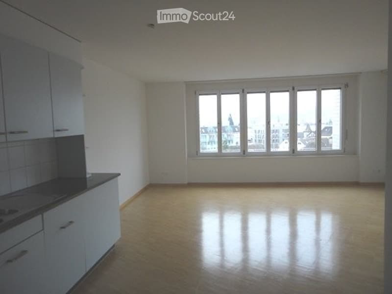Empty room with wooden flooring, white walls, and a fitted kitchen area with cabinets and a countertop. Large windows provide natural light and a view of the city.