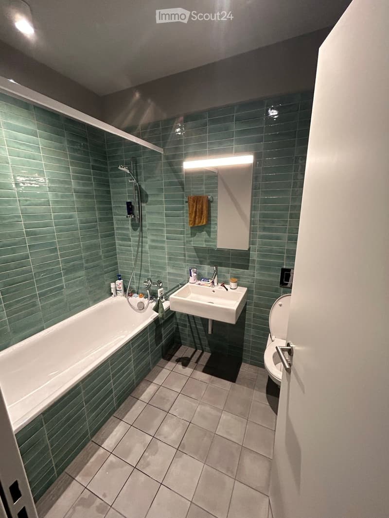 Modern bathroom with green tiled walls, a bathtub, a sink, a toilet, and a mirror with built-in lighting.