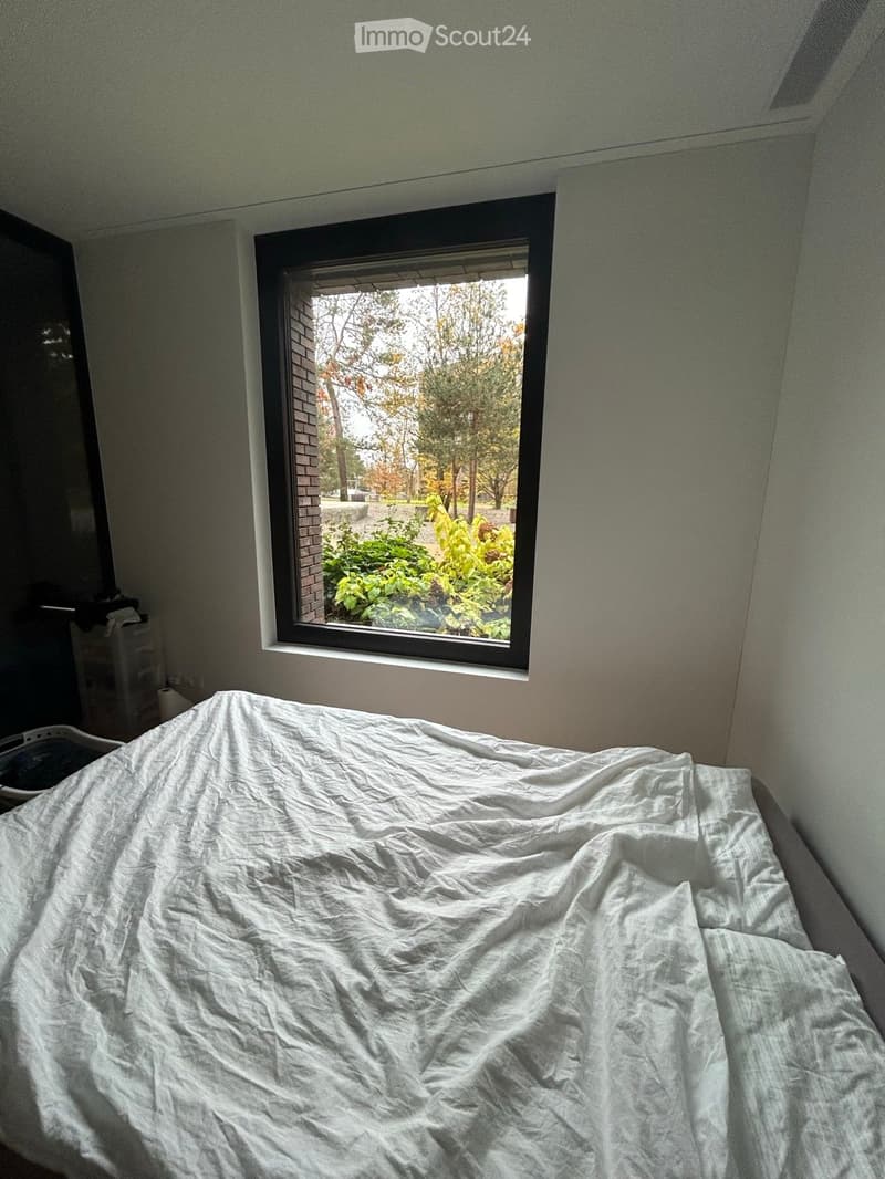 A bedroom with a single bed covered in a white sheet, a window showing a view of greenery and trees, and a small cabinet with items on it.