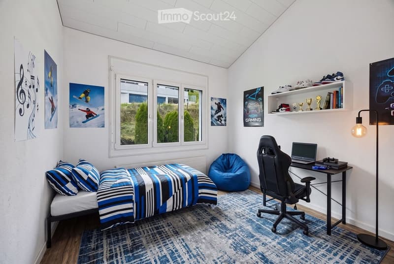 A modern bedroom with a single bed, a gaming chair, a laptop desk, a bean bag chair, and shelves with trophies and books. The room features large windows with a view of greenery outside.
