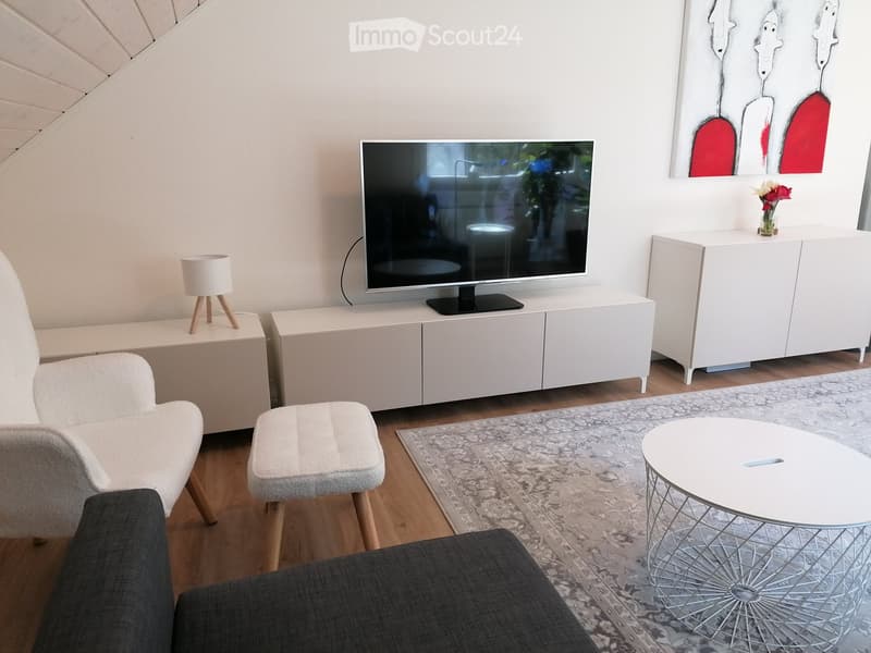 Modern living room with a grey sofa, armchair, small round table, TV on a white cabinet, and a decorative vase on a sideboard.