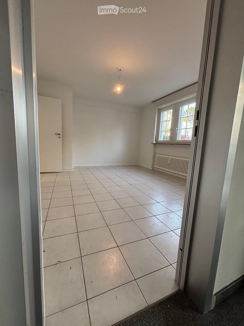 Empty room with tiled floor, white walls, a single window with a radiator below, and a door on the left side.