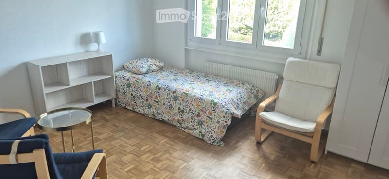 A small, well-lit bedroom with wooden flooring, a single bed with a floral duvet, a white shelf unit, a glass coffee table, and two chairs.