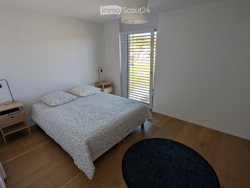 A modern bedroom with a wooden floor, a bed with a patterned duvet and pillows, a nightstand with a lamp, and a round stool near the window.