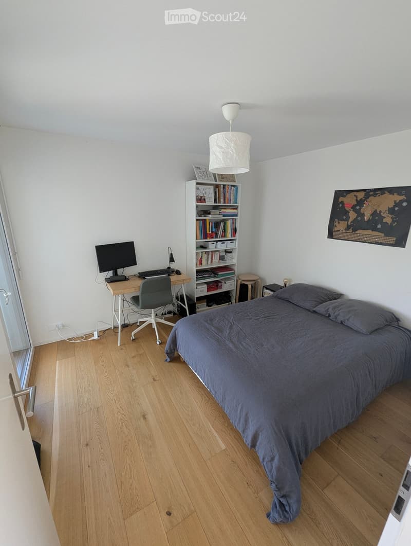 A modern bedroom with a large bed, a desk with a computer, a bookshelf, and a world map on the wall. The room features wooden flooring and white walls.