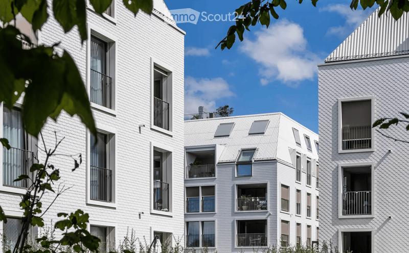 Modern white apartment building with multiple balconies and large windows, surrounded by greenery.