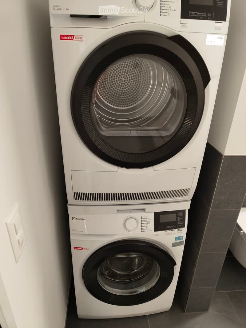 A modern bathroom with a white washing machine and dryer stacked vertically, featuring sleek design and control panels.