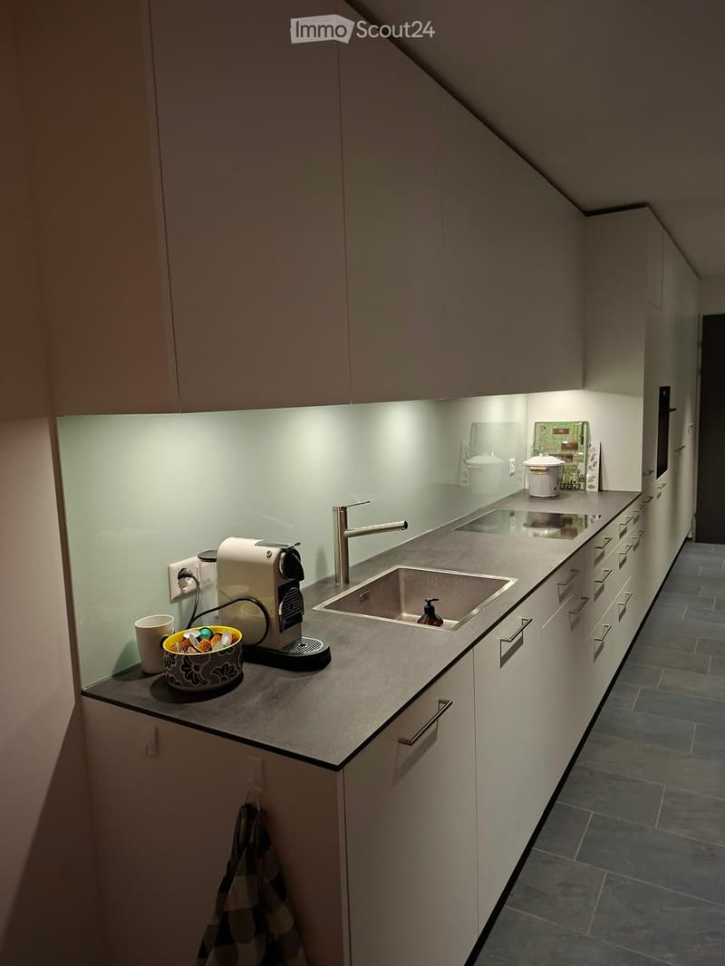 Modern kitchen with white cabinets, a stainless steel sink, and a coffee machine on the counter. The space features a sleek design with built-in lighting and a reflective backsplash.