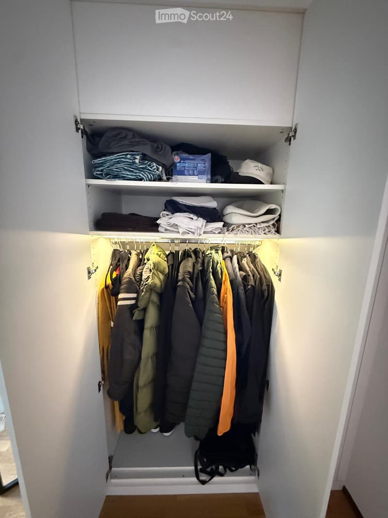 A well-organized closet with multiple shelves and a hanging rod, containing neatly arranged clothes, folded blankets, and a backpack on the floor.
