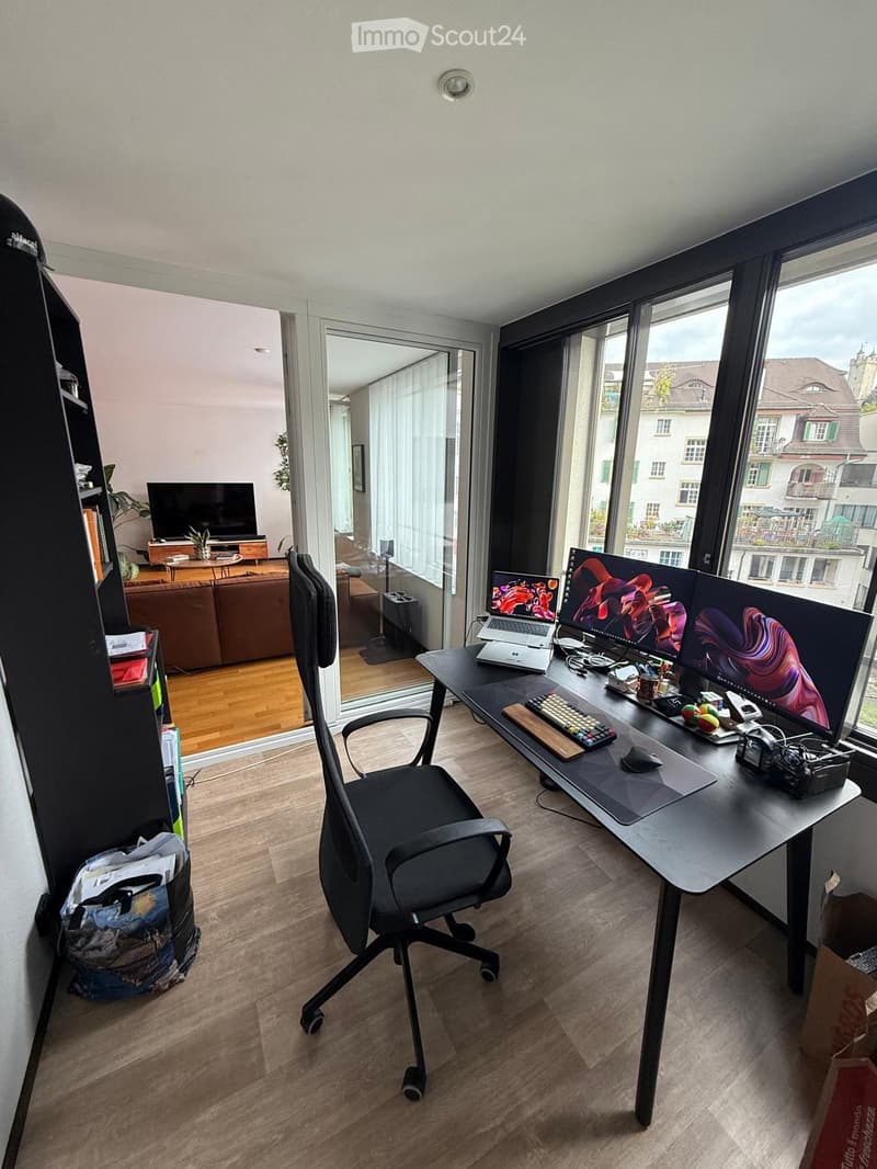 A modern, well-lit room with a large desk setup featuring a laptop, multiple monitors, and a comfortable office chair. The room has wooden flooring, large windows with a city view, and an open layout connecting to a living area with a sofa and TV.