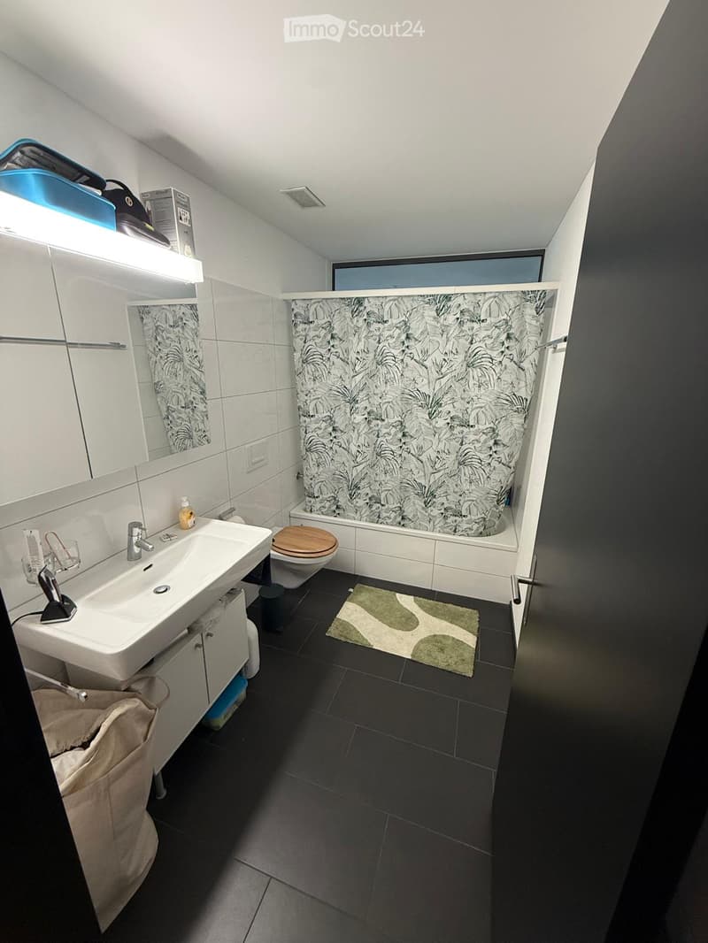 Modern bathroom with white tiles, dark floor, sink, toilet, bathtub with shower curtain, mirror, and small rug.