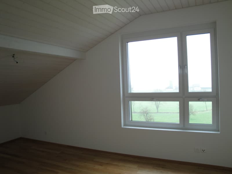 Empty room with wooden flooring, white walls, and large windows offering a view of a green landscape.