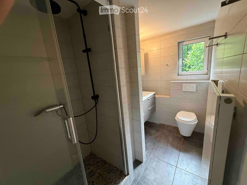 Modern bathroom with a walk-in shower, toilet, sink, and cabinet. Features tiled walls and a window with a view of greenery.