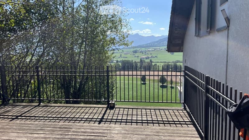 Wooden balcony with metal railing, overlooking a green field and distant mountains.