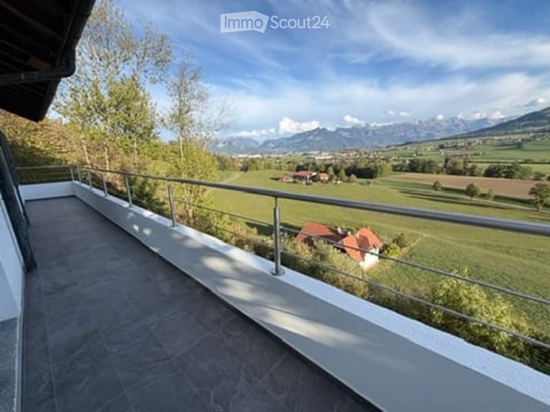 Balcony with metal railing overlooking a green field and distant mountains, with a house visible in the distance.