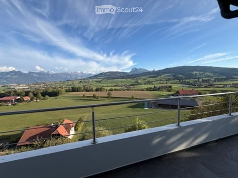 Balcony with metal railing overlooking a green field and distant mountains under a clear blue sky.