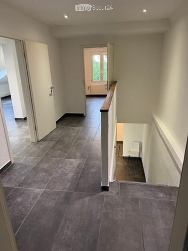 Spacious hallway with grey tiled flooring, white walls, and a wooden handrail leading to an upper level. Open doors reveal adjacent rooms.