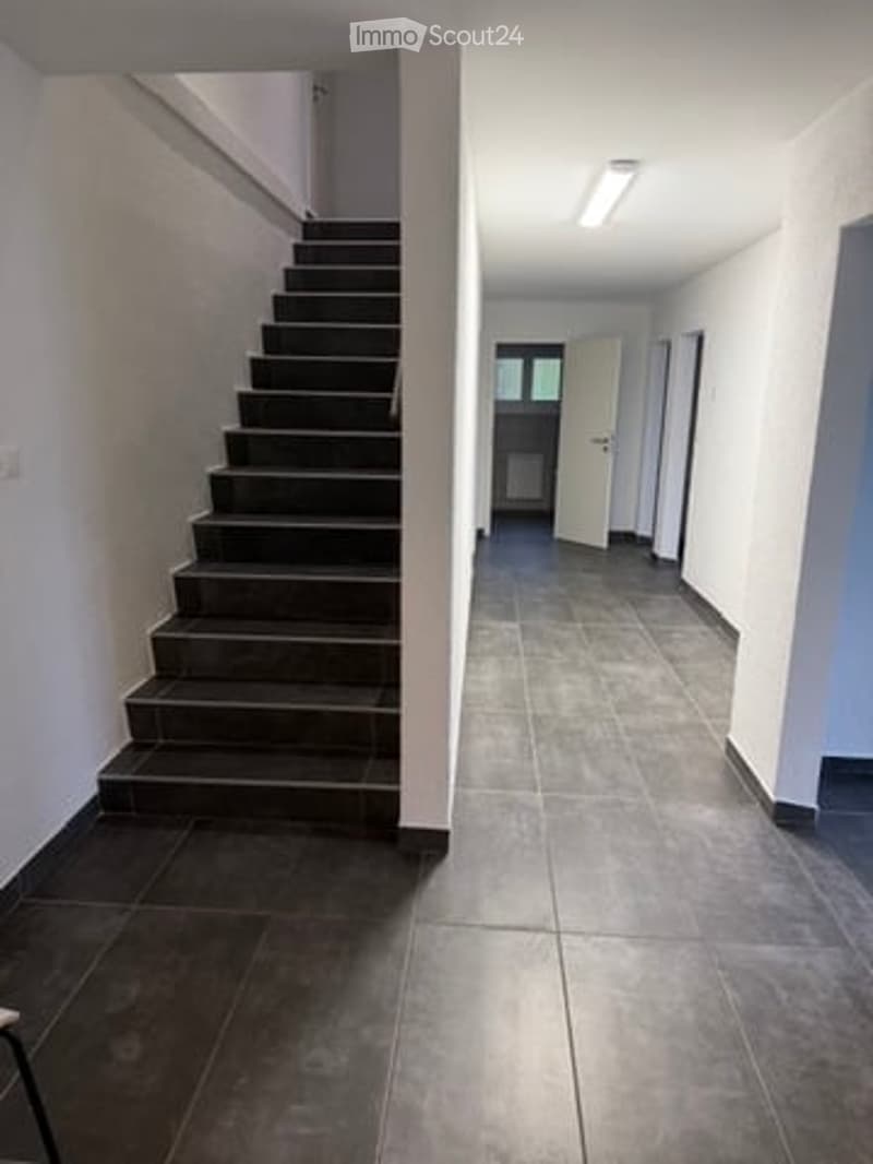 Empty hallway with tiled floor, white walls, and a staircase leading upwards. A door at the end is open, revealing another room.