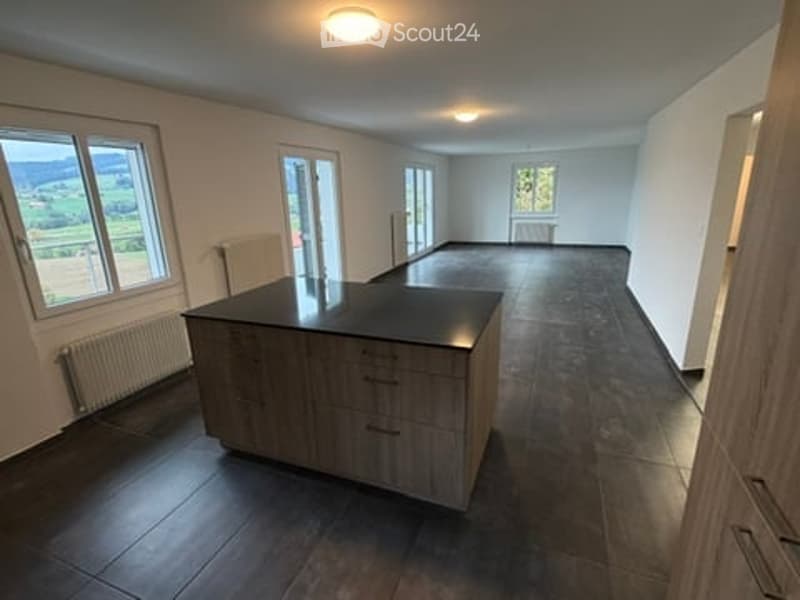 Modern kitchen with dark wooden floor, large windows offering a scenic view, kitchen island with cabinets, and radiators along the walls.