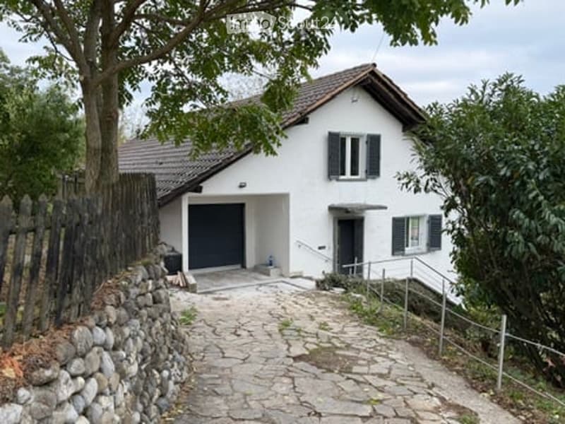 Single-story white house with a tiled roof, black shutters, and a garage. A stone pathway leads to the entrance, flanked by a wooden fence and greenery.