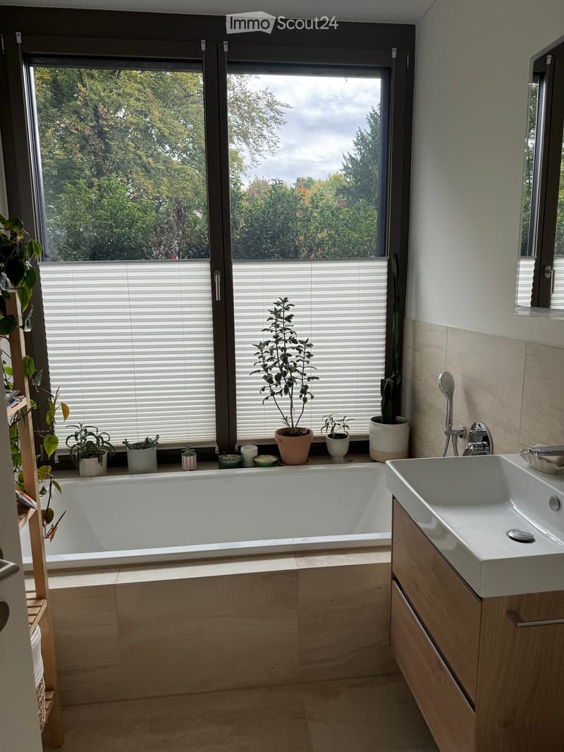 Modern bathroom with a large window, bathtub, sink, and several potted plants on the windowsill.