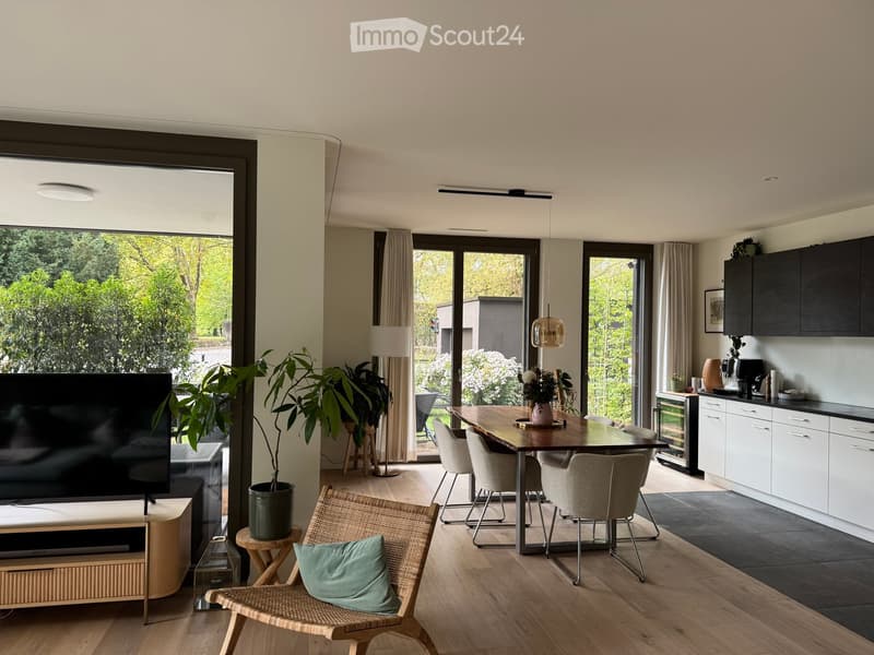 Open-plan living and dining area with modern furniture, large windows, and a kitchen area with cabinets and appliances.