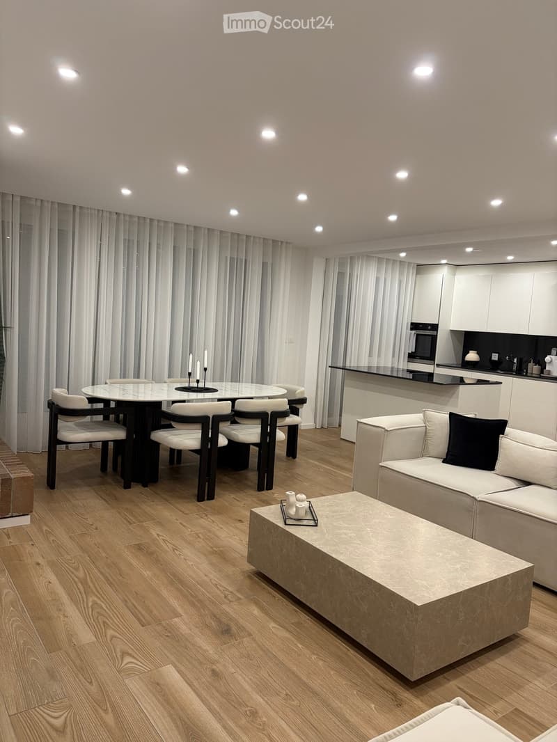 Modern living and dining area with a white sofa, coffee table, dining table with chairs, and a sleek kitchen area with white cabinets and an oven.
