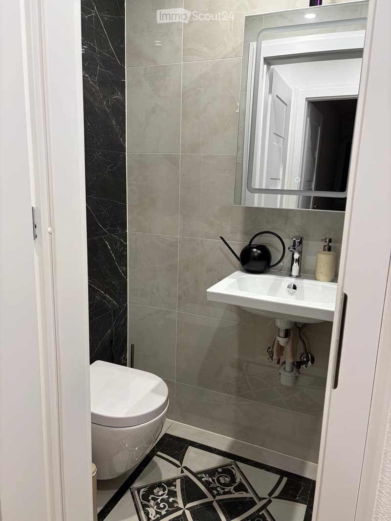 Modern bathroom with black and grey marble tiles, white toilet, sink, and mirror. Includes a decorative black teapot on the sink counter.