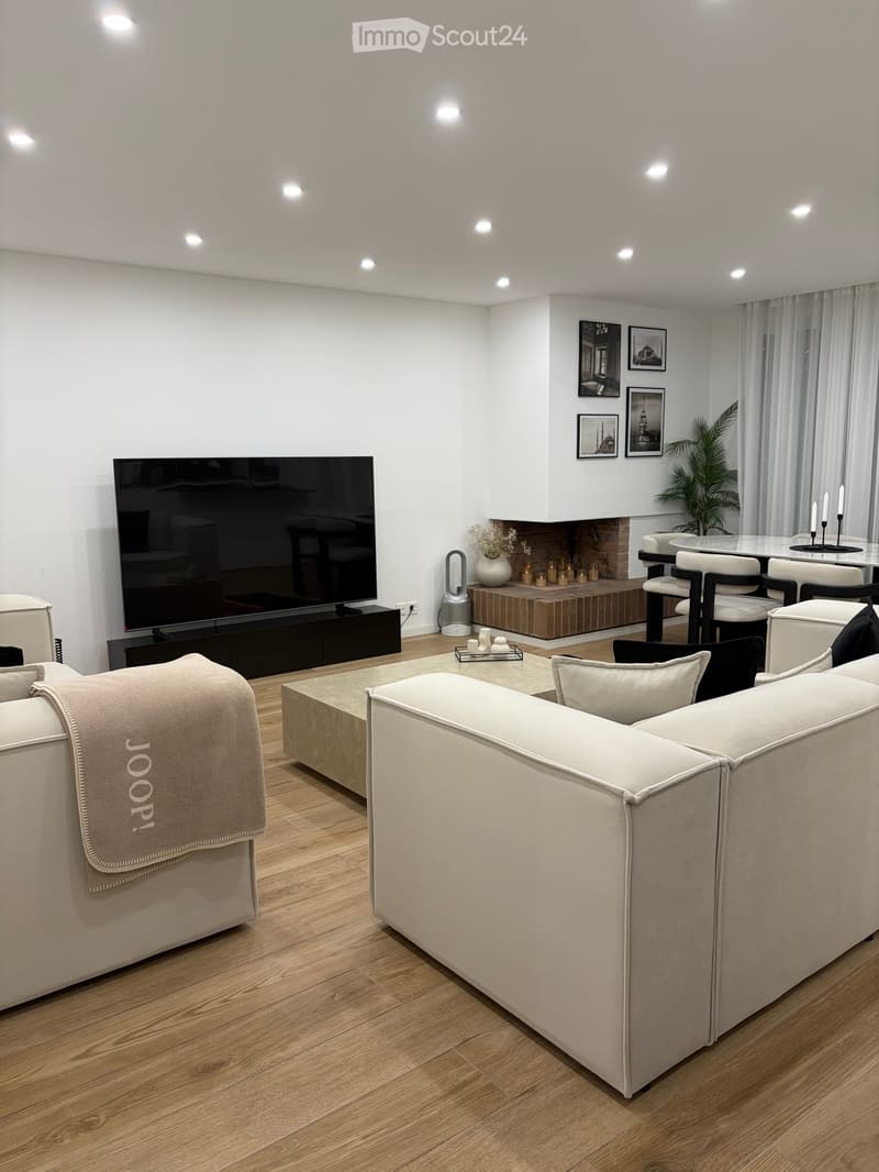 Modern living room with a large sectional sofa, coffee table, flat-screen TV, fireplace, dining area with chairs and table, and decorative plant and candles.