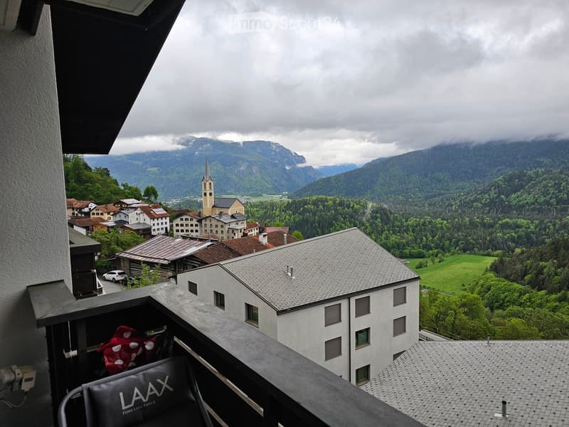 Balcony overlooking a picturesque village with traditional houses, a church tower, and lush green mountains under a cloudy sky.