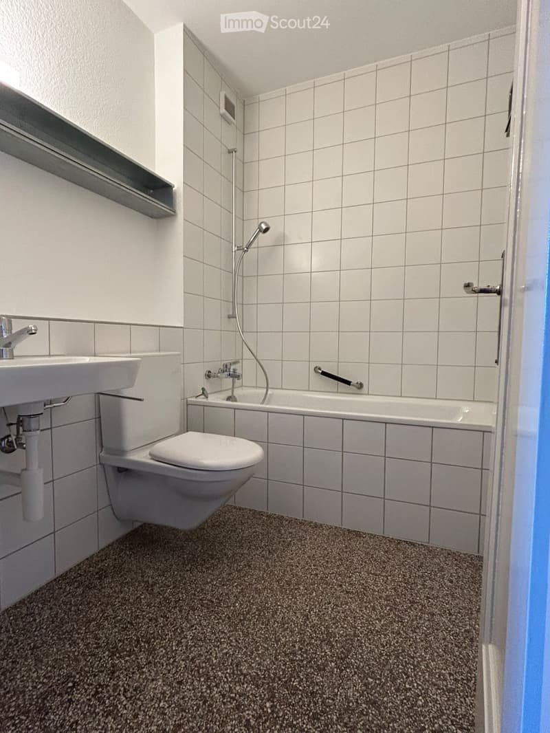 Modern bathroom with white tiled walls, a toilet, a sink, a bathtub with a showerhead, and a grey shelf above the sink.