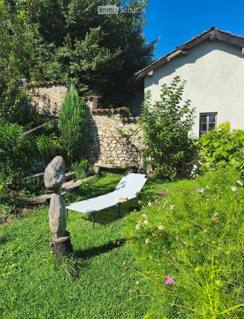 Garden with a white lounge chair, a wooden bench, a stone sculpture on a tree stump, and a stone wall with a small white house in the background.