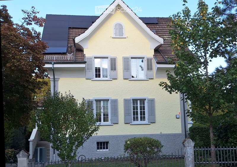 Two-story yellow house with white shutters, multiple windows, and a small balcony on the upper floor. The house features a gray stone base and is surrounded by trees and a metal fence.