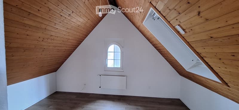 Empty attic room with wooden ceiling, white walls, wooden floor, and a small window with a white frame.