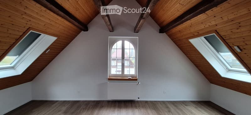 Empty attic room with wooden ceiling beams, wooden flooring, and multiple windows including a central arched window and two skylights.